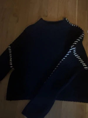 Svart stickad tröja med vita detaljer - Säljer en svart stickad tröja med lång ärm och coola vita snörningsdetaljer längs axlar och ärmar. Tröjan har en grovstickad struktur och en klassisk rund hals. Endast använd nån gång! Som helt ny! 