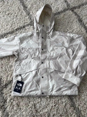 The North Face x KAWS camo vindjacka - Säljer en limited edition vindjacka från The North Face x KAWS i ljusgrått och vitt camo-mönster. Jackan har huva med snörning, tryckknappar framtill, DryVent-material och broderade XX-detaljer. Perfekt för dig som gillar streetwear och unika samarbeten.