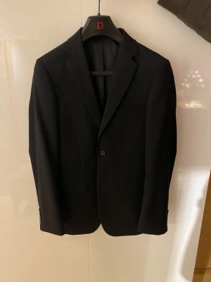 Svart kostym Dressmann slim fit - Säljer en svart tvådelad kostym från Dressmann i slim fit-modell. Klassisk kavaj med två knappar och matchande byxor. Snygg passform och tidlös look, perfekt för dig som vill vara stilsäker. Storlek på kavaj är 42 Regular och byxa 28/32. Använd endast en gång!