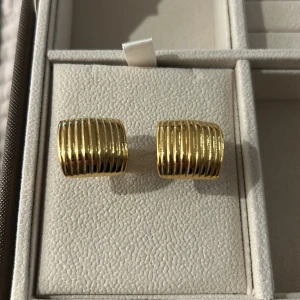 Guldiga ribbade örhängen från Muli collection  - Säljer ett par chunky örhängen i guldig metall med ribbad, räfflad yta. De har en fyrkantig, lätt böjd form som sitter snyggt på örat och ger en cool statement-look. Perfekta för att lyfta vilken outfit som helst. Ordinarie 599kr pris 400kr