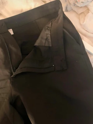 Svarta kostymbyxor med dragkedja - Snygga svarta kostymbyxor med klassisk passform och dragkedja framtill. Byxorna har hällor för bälte och sidfickor, perfekta för en stilren look. Materialet känns slätt och dressat, passar till skjorta eller t-shirt.