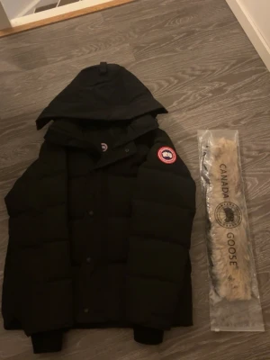 Canada Goose wyndham red Label - Svart dunjacka från Canada Goose med stor huva och klassisk logga på ärmen. Jackan har en puffer-design, ribbade muddar och medföljande pälsdetalj till huvan. Perfekt för kalla dagar och har en clean, stilren look. Skriv vid frågor eller prisförslag.