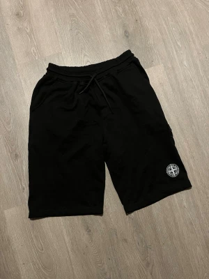 Svarta shorts från Stone Island - Svarta shorts från Stone Island med klassisk logotyp på benet. De har elastisk midja med snörning, dragkedjeficka och en loose passform. Perfekta för chill dagar och streetwear-stil. Materialet är mjuk bomull som känns skönt mot huden.