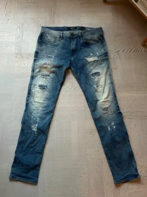 Replay Anbass - Säljer ett par Replay Anbass jeans i blå tvätt med slitna detaljer och distressed look. Jeansen har klassisk femficksmodell, normal midja och raka ben. Materialet är mjukt jeans-tyg som ger en avslappnad vibe. Hör av dig om frågor!.