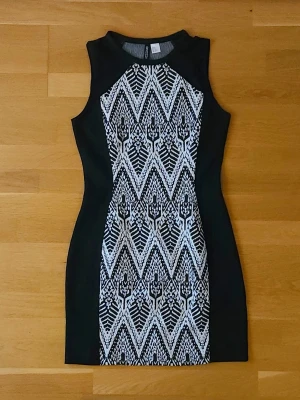 Svartvit klänning - Svartvit kort klänning från H&M med grafiskt mönster framtill. Klänningen är ärmlös, har rund halsringning och figurnära passform. Sidorna är helsvarta vilket ger en snygg siluett. Perfekt för dig som gillar statement-plagg!