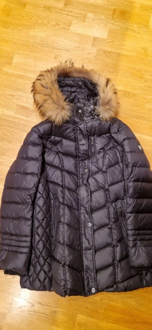 SAKI Sweden svart dunjacka med fuskpäls, avtagbar huva - Snygg svart dunjacka med fluffig fuskpäls på huvan. Jackan har quiltade detaljer på sidorna och flera dragkedjor framtill. Innerficka. Mycket fint skick, från rök- och djurfritt hem. Nypris ca 3000 kr.