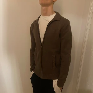 Brun stickad kofta med dragkedja - Snygg brun stickad kofta med dragkedja framtill och ribbade detaljer vid krage, muddar och fickor. Koftan har långärmad passform och är perfekt för lager-på-lager. Enkel och stilren design som funkar till många olika looks. Om du köper använd PostNord eller instabox helst!