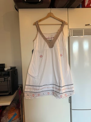 Odd Molly vit kortklänning med spets - Supersöt vit klänning med beige spetsdetaljer vid v-ringningen och axelbanden. Broderade blommor i rosa och gult samt dekorativa sömmar nertill ger en bohemisk vibe. Klänningen är ärmlös och har en luftig passform, perfekt för varma dagar. Från märket Odd Molly. Storlek 2 som enligt deras storleksguide motsvarar en M. Mycket fint skick, jag hittar inga synliga anmärkningar eller defekter. 