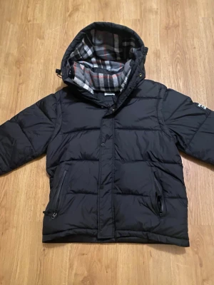 Svart pufferjacka från Burberry - Svart pufferjacka från Burberry med huva och rutigt foder i grått, vitt och rött. Jackan har dragkedjor på fickorna och en stor Burberry London England-patch på ärmen. 