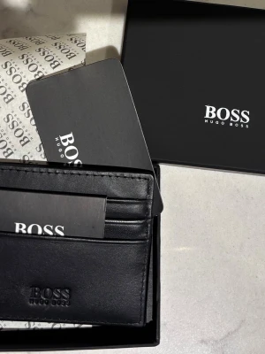 Svart kortfodral från Hugo Boss - Helt ny svart kortfodral från Hugo Boss i skinn. Fodralet har flera kortfack och diskret logga framtill. Kommer i originalask med Boss-tryck, perfekt för dig som vill ha en clean och lyxig accessoar till dina kort. Se bild för jämförelse med ipod brevid. Skickar snabbt! 