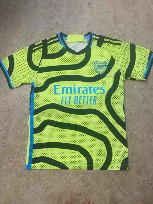 Arsenal neon gul matchtröja - Säljer en Arsenal matchtröja i neon gul med svarta vågiga ränder och små svarta prickar. Tröjan har blå detaljer vid krage och ärmslut samt klubbmärke på bröstet. Materialet är lätt och andas, perfekt för fotbollsträning eller att bära på läktaren.