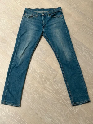 Levi's 511 blå jeans W31 L32 - Säljer ett par Levi's 511 jeans i klassisk blå tvätt med snygga slitningar. Modellen är slim och har raka ben, fem fickor och detaljerade sömmar. Jeansen är tillverkade i bomull och har en läderpatch bak i midjan. Perfekt passform för dig som gillar en modern look. Den finns en väldigt liten fläck med blå målarfärg på vänstra benet.
