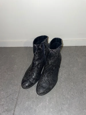 Svarta boots med ormskinnsmönster från Ecco - Snygga svarta boots från Ecco i storlek 36 med ormskinnsmönster och spetsig tå. Skorna har låg klack och dragkedja på sidan för enkel på- och avtagning. Tillverkade i skinn med en cool, mörk finish som ger en edgy vibe.