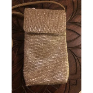 Glittrig guld axelväska med lock (passar telefon) - Snygg liten axelväska i glittrigt guld med rektangulär form och lock med magnetknäppning. Perfekt storlek för mobilen och småsaker. Väskan har ett smalt axelband och insidan är fodrad i beige tyg. Riktigt glammig och sticker ut med sitt glittriga material.