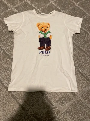 Vit Polo Ralph Lauren t-shirt med björn - Säljer en fet Ralph Lauren t shirt för bra pris. Storlek 12-13 år. Säljer för jag växte ut ur tröjan, en av mina favorit tröjor, skriv om ni vill ha information eller fler bilder!