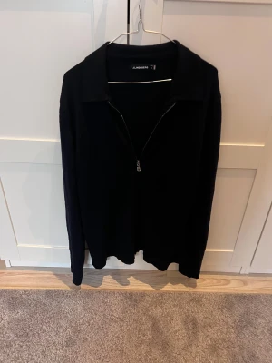 Svart half zip tröja från J.Lindeberg - Svart långärmad tröja från J.Lindeberg med krage och half zip-dragkedja framtill. Stilren och enkel design som funkar till många olika looks. Materialet känns mjukt och skönt mot huden, perfekt för dig som gillar klassiska plagg med en modern touch.