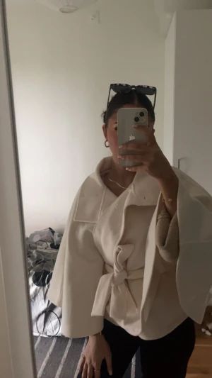 Beige capejacka med bälte Gina Tricot - Säljer en beige capejacka från Gina Tricot med bred krage, knappar och knytbälte i midjan. Jackan har en loose passform och vida ärmar som ger en cool och modern siluett. Perfekt för dig som gillar stilrena och trendiga plagg.