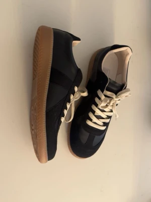 Svarta maison margiela gats skor - Säljer ett par svarta maison margiela (helt nya aldrig använda) sneakers med beige snörning och gummisula i brunt. Skorna har en snygg mix av skinn och mocka på ovandelen, rund tå och platt sula. Insidan är ljus och mjuk för extra komfort. Perfekt för dig som gillar stilrena och klassiska sneakers. Storleken är 44 men passar mindre som 42-43
