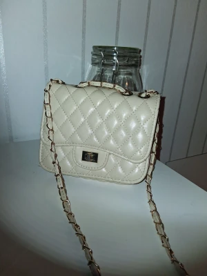 Beige quiltad axelväska med kedja - Snygg beige axelväska med quiltad design och guldfärgad kedja som axelrem. Väskan har ett lock med vridlås och insidan har ett fack med dragkedja. Perfekt storlek för det viktigaste och ger en chic vibe till din outfit.