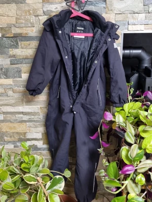 Svart overall från Reima med fuskpäls - Svart täckoverall från Reima med avtagbar huva och fuskpäls. Overallen har dragkedja framtill, reflexdetaljer och resår vid ärm- och benslut. Perfekt för kalla vinterdagar och håller dig varm och torr. Snygg och funktionell design för aktiva dagar utomhus. 