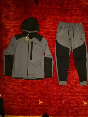 Nike Tech Fleece Tracksuit - Nike Tech Fleece tracksuit – helt ny med prislapp! Snygg tvådelad set i grått och svart, består av hoodie med dragkedja och matchande byxor. Aldrig använd, fortfarande med tag kvar. Perfekt passform, varm och lätt – klassisk Nike-kvalitet.