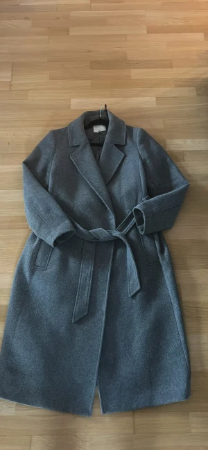Grå kappa med bälte från H&M - Snygg grå kappa från H&M i storlek 40. Klassisk modell med bred krage, knytskärp i midjan och diskreta fickor på sidorna. Perfekt för dig som vill ha en stilren och tidlös look under kyliga dagar.
