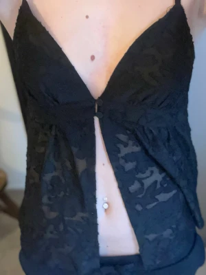 Svart spetsig linnetopp med knappar - Säljer en svart linnetopp med tunna axelband och v-ringning. Toppen är i transparent spets med blommigt mönster och har två knappar framtill. Den är croppad och har en luftig passform som ger en snygg och trendig look.