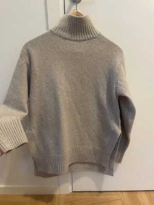 Beige stickad polotröja med ribbad krage - Snygg beige stickad polotröja med bred ribbad krage och muddar. Tröjan har en loose passform och är perfekt för kyliga dagar. Materialet är mjukt och värmande, och den neutrala färgen gör den enkel att matcha med allt. Klassisk design med slits i sidorna och diskreta detaljer.