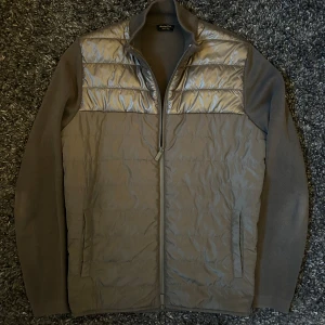 Grå quiltad pufferjacka från Massimo Dutti - Snygg grå pufferjacka från Massimo Dutti med quiltade paneler och stickade ärmar. Jackan har hel dragkedja framtill, ståkrage och två sidofickor. Materialmix med glansig syntet på kroppen och mjuka stickade partier på ärmarna. Perfekt för dig som gillar stilren och modern look.