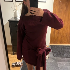 Vinröd stickad klänning med rosett - Supermysig vinröd stickad klänning med lång ärm och stor rosett i midjan. Modellen är kort och har en loose passform med breda ärmslut. Perfekt för dig som gillar mjuka material och vill ha en enkel men snygg look.