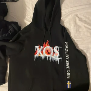 Svart XQS hoodie med flames och is - SKRIV OM NI VILL DISKUTERA PRISET!!! Svart hoodie från XQS med stor logga framtill i eld- och isdesign. Texten 'MADE BY SWEDEN' och svensk flagga på ena ärmen. Klassisk huva och känguruficka. Perfekt statement-plagg för dig som gillar streetwear. Den är sprillans ny så!