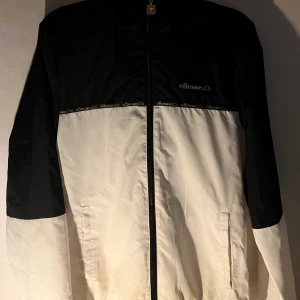 vindjacka från Ellesse - Svartvit vindjacka från Ellesse i storlek XS. Jackan har huva, dragkedja med Ellesse-logga och logoband över bröstet. Långärmad och med två fickor framtill. Perfekt för dig som gillar sportig och clean stil. Den är ny