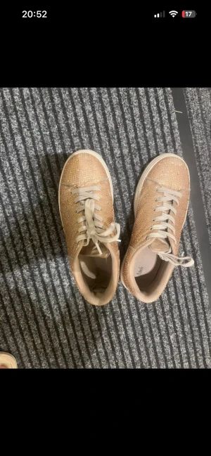 Glittriga beige sneakers med snörning - Säljer ett par snygga sneakers i beige med glittriga detaljer över hela skon. De har rund tå, platt vit sula och klassisk snörning. Perfekta för dig som vill sticka ut med lite extra bling i vardagen. Skorna har en modern och ungdomlig look.