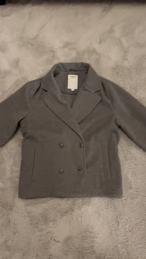 Grå skepparkavaj från Kingsmile - Snygg grå skepparkavaj från Kingsmile med klassisk dubbelknäppning och breda slag. Jackan har två framfickor och är tillverkad i mjukt material som ger en stilren look. Perfekt för dig som gillar minimalistisk och tidlös stil.