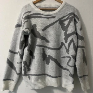 Vit och grå stickad tröja med mönster - Säljer en vit stickad tröja med grått abstrakt mönster. Tröjan har rund hals, ribbade muddar och långärmad passform. Den är loose och har en skön vibe som passar perfekt när du vill vara chill men ändå se cool ut.