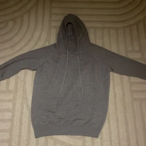 Grå hoodie med huva och snörning - En enkel grå hoodie med huva och långa ärmar. Tröjan har snörning vid halsen och ribbade muddar vid ärmslut och nederkant. Materialet känns mjukt och stickat, perfekt för chill dagar. Passformen är normal och färgen är mörkgrå.