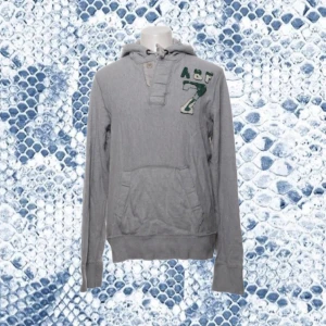 Vintage hoodie Abercrombie & Fitch  - Snygg grå hoodie från Abercrombie & Fitch i storlek L men sitter som M. Tröjan har huva, magficka och broderad A&F-logga med siffran 7 på bröstet. Tillverkad i mjuk bomull, perfekt för chill dagar. Långärmad med knappdetalj vid halsen.