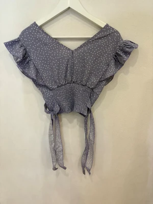 Ljuslila prickig omlottopp från Shein - Supersöt ljuslila omlottopp från Shein med vita prickar, volangdetaljer på axlarna och knytband i midjan. Toppen har v-ringning och smockad midja som ger en snygg passform. Perfekt för dig som gillar en feminin och trendig stil.