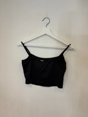 Svart croppad linnetopp med smala band - Snygg svart croppad linnetopp med tunna axelband och enkel design. Toppen har en lätt glansig finish och bredare kant nedtill för en stilren look. Perfekt att matcha med jeans eller kjol för en trendig outfit.