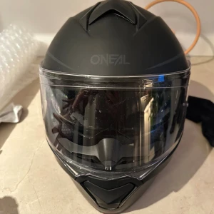Svart O'Neal MC-hjälm med visir - Snygg svart MC-hjälm från O'Neal med matt finish och integrerat klart visir samt inbyggt solvisir. Hjälmen har aerodynamisk form och ventiler för extra komfort. Insidan är vadderad med mesh-tyg för skön passform. Perfekt för dig som vill ha både stil och säkerhet på vägen. Nyskick använd 3ggr 