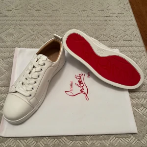 Christian Louboutin vita sneakers - Snygga vita sneakers från Christian Louboutin med ikonisk röd sula och diskret logga. Skorna har rund tå, snörning och är tillverkade i lyxigt skinn. Perfekt för dig som vill sticka ut med stilrena och exklusiva sneakers.