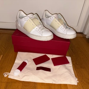 Valentino Open  - Säljer ett par Valentino Open sneakers i vitt skinn med en bred ljusbeige rem över mitten. Klassisk rund tå, platt sula och snörning framtill. Snygga detaljer med nitar på hälen och diskret logga på plösen. Og saker följer med vid snabb och smidig affär 