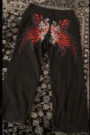 Svarta jeans med röd-vitt tryck bak - Svarta jeans med rak passform och ett stort, iögonfallande rödvitt grafiskt tryck över bakfickorna. Klassisk femficksmodell med bälteshällor och knappgylf. Materialet är jeans och byxorna har en cool streetkänsla.priset går att diskuteras vid en snabb äffar