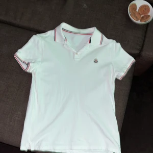 Vit Moncler pikétröja med ränder - Vit pikétröja från Moncler med klassisk krage och korta ärmar. Tröjan har snygga rödblå ränder på krage och ärmslut samt Moncler-logga på bröstet. Tillverkad i bomull för skön känsla och stilren look. Perfekt för dig som gillar exklusiva märken och enkel design. Skanna QR koden så ser ni att den är äkta