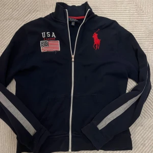 Mörkblå zipjacka Polo Ralph Lauren XL - Mörkblå zipjacka från Polo Ralph Lauren i storlek XL med gråa ränder längs ärmarna, broderad röd logga på bröstet och USA-flagga med text. Jackan har ståkrage, två fickor fram och hel dragkedja. Perfekt för en sportig och chill stil. Obs tröjans storlek ärnsom storlek M