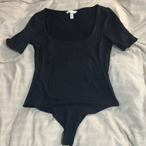 Svart bodysuit med kort ärm från H&M - En svart bodysuit från H&M med djup rundad urringning och korta ärmar. Gjord i mjukt stretchigt material som sitter tight mot kroppen. Perfekt att styla med jeans eller kjol för en clean look.