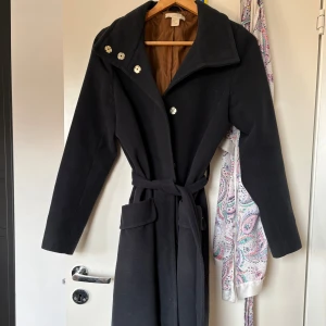 Svart kappa med bälte och blommiga knappar - Svart kappa från H&M med snyggt bälte i midjan och blommiga knappar framtill. Jackan har bred krage, två fickor och är helfodrad med brunt innerfoder. Perfekt för dig som gillar stilrena och klassiska plagg med en twist.