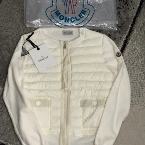 Vit quiltad cardigan jacka Moncler - Snygg vit cardigan jacka från Moncler med quiltad framsida och två fickor med knappdetaljer. Jackan har rund hals, dragkedja och Moncler-logga på ärmen. Materialmix av dun och stickad bomull ger en cool och lyxig vibe.💕