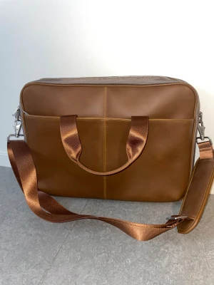 Brun datorväska från Sandstrøm - Stilren datorväska i brunt skinn från Sandstrøm med avtagbar axelrem och praktiska handtag. Väskan har ett rymligt, vadderat innerfack och stängs med dragkedja. Perfekt för att bära laptop och tillbehör på ett snyggt sätt.