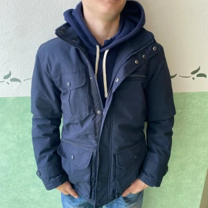 Mörkblå field jacket med fickor - Parkajacka i mörkblå färg med flera stora fickor framtill, dragkedja och tryckknappar. Jackan har hög krage med detaljerade metallknappar och är långärmad. Materialet är slitstark polyester, perfekt för kyliga dagar.